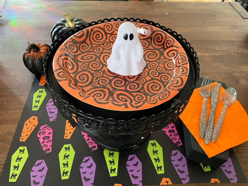May include: A black and orange Halloween themed paper plate with a white ghost on top. The plate is on a black metal stand. The stand is on a black and green table runner with a pattern of coffins and bats.