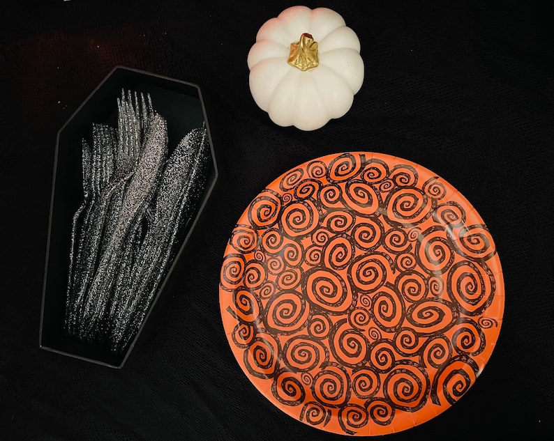 May include: A black coffin-shaped container filled with silver plastic cutlery. A white pumpkin with a gold stem sits on a black surface next to a bright orange paper plate with a black swirl pattern.