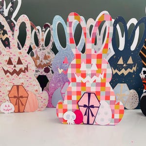 May include: A collection of decorative bunny-shaped cutouts with jack-o'-lantern faces and gift box accents. The cutouts feature various patterns and colors, including floral, plaid, and solid hues. A white pumpkin with a gold stem sits in front.