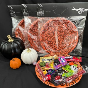 May include: A package of three orange and black paper plates with a swirling pattern. The plates are in a clear plastic bag with a black header that says "Skelly Paper Co.". The package is on a black surface with three small pumpkins and a plate of Halloween candy.