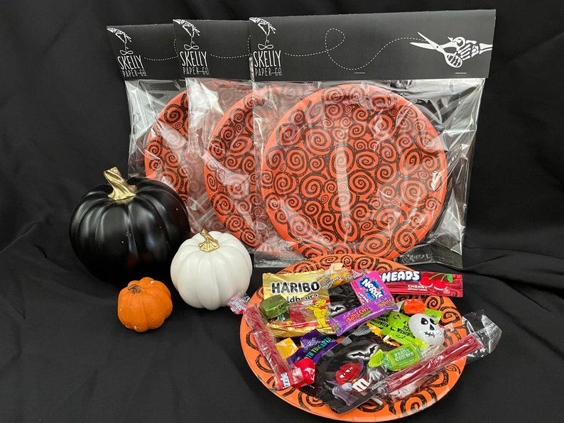 May include: A package of three orange and black paper plates with a swirling pattern. The plates are in a clear plastic bag with a black header that says "Skelly Paper Co.". The package is on a black surface with three small pumpkins and a plate of Halloween candy.