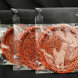 May include: Three orange paper plates with a black swirl pattern. The plates are packaged in clear plastic bags with a black header that reads "Skelly Paper-Co."