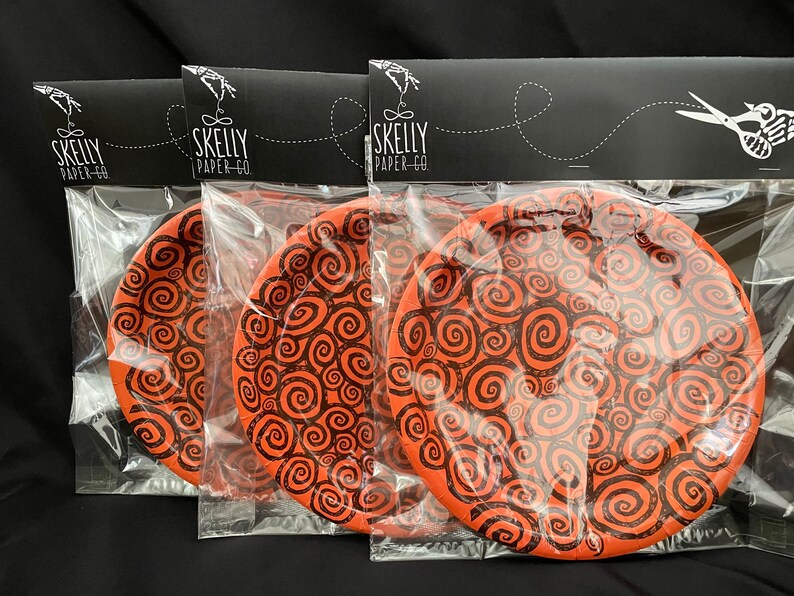 May include: Three orange paper plates with a black swirl pattern. The plates are packaged in clear plastic bags with a black header that reads "Skelly Paper-Co."