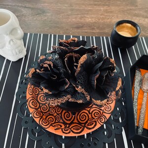 May include: A black and orange Halloween table setting with a black and gold flower centerpiece, a white skull mug, a black cauldron, and silver cutlery on a black and white striped placemat.