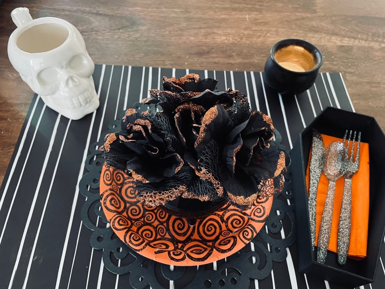 May include: A black and orange Halloween table setting with a black and gold flower centerpiece, a white skull mug, a black cauldron, and silver cutlery on a black and white striped placemat.