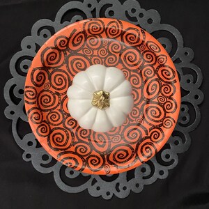 May include: A white ceramic pumpkin with a gold stem sits on top of an orange paper plate with a black swirl pattern. The plate is on a black lace doily.