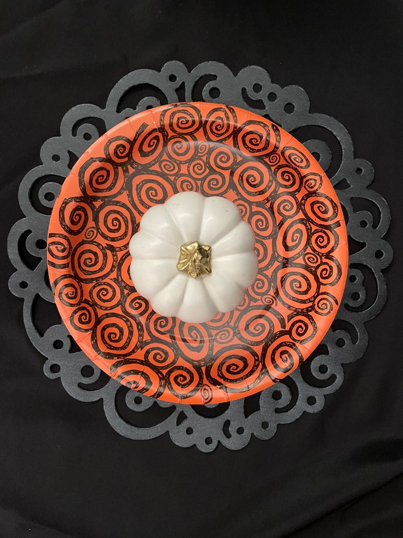 May include: A white ceramic pumpkin with a gold stem sits on top of an orange paper plate with a black swirl pattern. The plate is on a black lace doily.