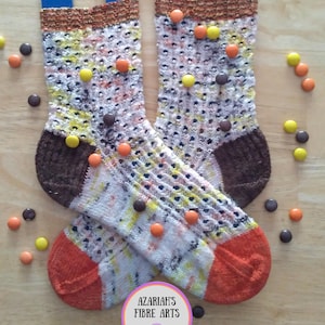 May include: A pair of hand-knitted socks featuring a speckled pattern in white, yellow, orange, and dark brown, with solid brown heels and orange toes. Small round candies in yellow, orange, and brown are scattered over the socks and surface.