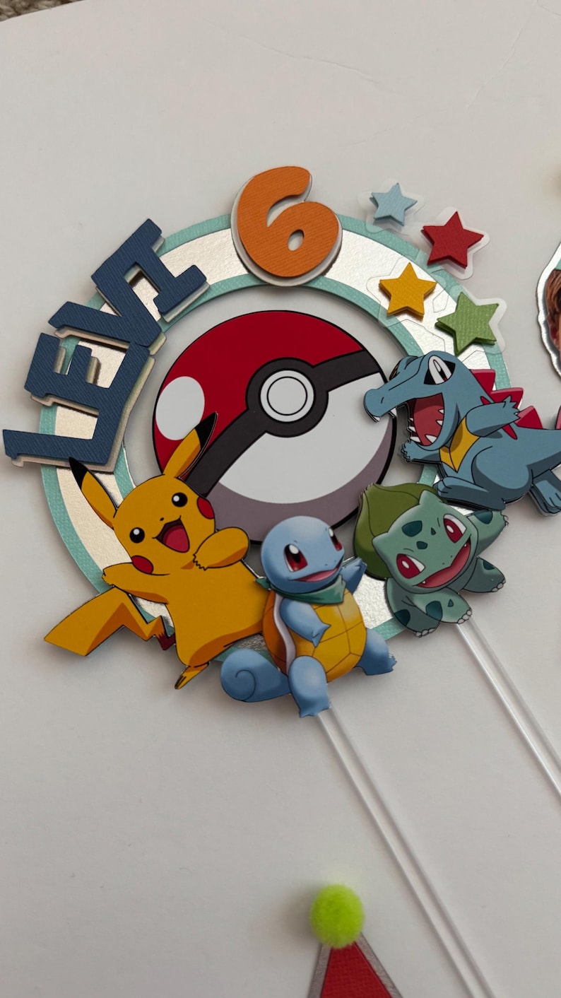 Pu&ograve; includere: Un topper per torta con una Pok&eacute; Ball rossa e bianca, un Pikachu giallo, uno Squirtle blu, un Bulbasaur verde e un Charmander blu e verde. Il topper include anche il nome "Levi" e il numero "6" in arancione.