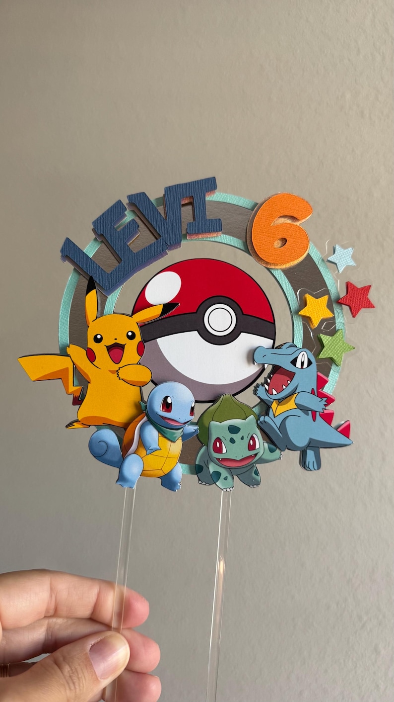Pu&ograve; includere: Un topper per torta con un design di personaggi Pokemon blu, verde e giallo. Il topper include una Pok&eacute;ball, il numero 6 e il nome "Levi".