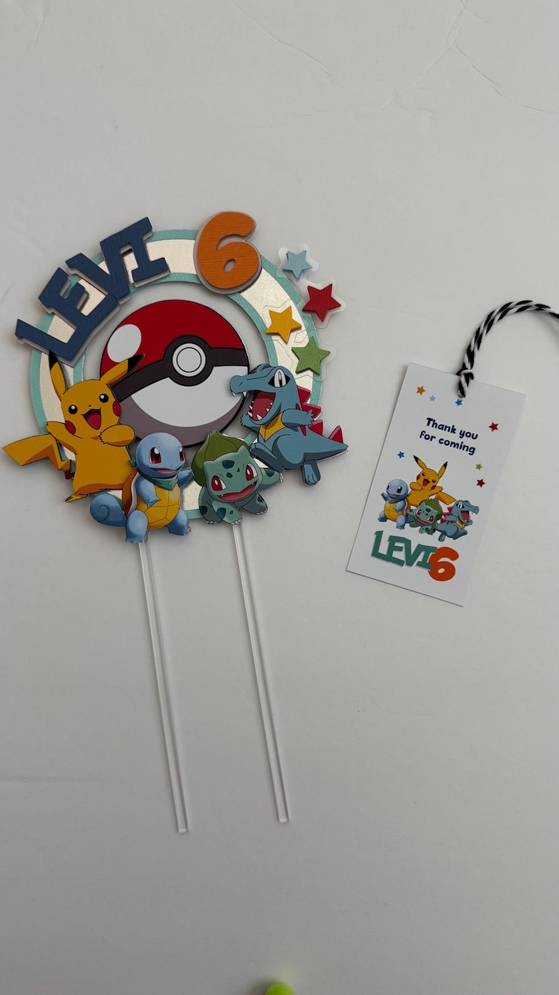 Pu&ograve; includere: Un topper per torta a tema Pok&eacute;mon con Pikachu, Bulbasaur, Squirtle e Charmander. Il topper include una Pok&eacute; Ball rossa e bianca, un cerchio blu e verde con il nome "Levi" e il numero "6" in arancione. Un'etichetta separata dice "Grazie per essere venuto" con un'immagine di Pok&eacute;mon e "Levi 6".