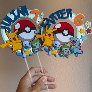 Pu&ograve; includere: Due topper per torte a tema Pok&eacute;mon con i nomi "Julian 7" e "Carter 6" in lettere blu su uno sfondo verde e arancione. Ogni topper presenta un Pikachu, una Pok&eacute; Ball e tre personaggi Pok&eacute;mon.