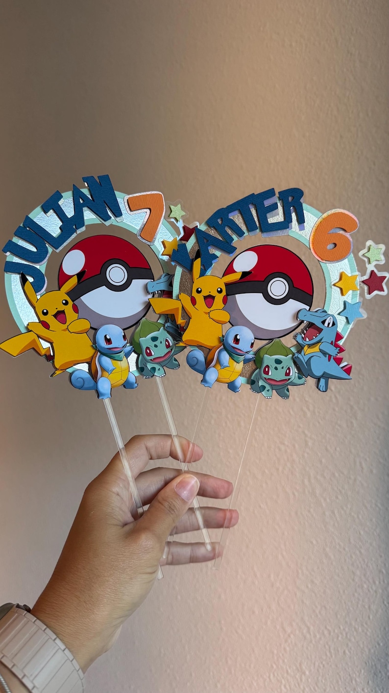 Pu&ograve; includere: Due topper per torte a tema Pok&eacute;mon con i nomi "Julian 7" e "Carter 6" in lettere blu su uno sfondo verde e arancione. Ogni topper presenta un Pikachu, una Pok&eacute; Ball e tre personaggi Pok&eacute;mon.