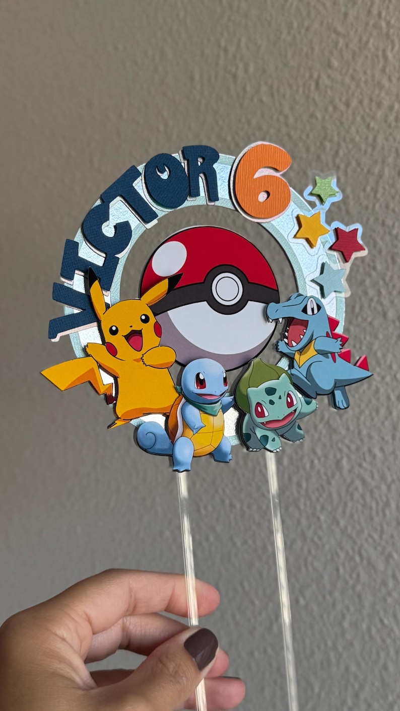 Pu&ograve; includere: Un topper per torta con una Pok&eacute; Ball blu, rossa e bianca con un Pikachu giallo, uno Squirtle blu e un Bulbasaur verde. Il topper include anche il nome "Victor" e il numero "6" in un cerchio blu con stelle arancioni.