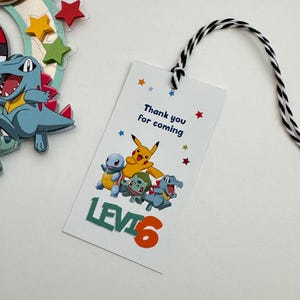 Pu&ograve; includere: Un'etichetta di ringraziamento bianca con un'immagine di personaggi Pokemon in cartone animato colorato. L'etichetta dice "Grazie per essere venuto" e "Livello 6".