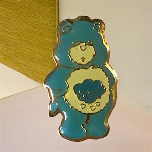 May include: A blue Care Bear badge with a gold border. The bear features a yellow tummy with a blue cloud design. A collectible item.