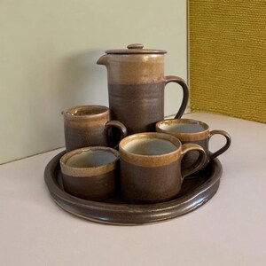 May include: A brown ceramic coffee set comprising a coffee pot with lid, four mugs, and a serving tray. The mugs and coffee pot feature a two-tone design with a lighter brown rim. The set is designed for serving coffee or tea.