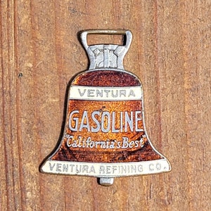 May include: A silver bell-shaped keychain with a brown and white enamel design. The keychain says "Ventura Gasoline California's Best Ventura Refining Co."