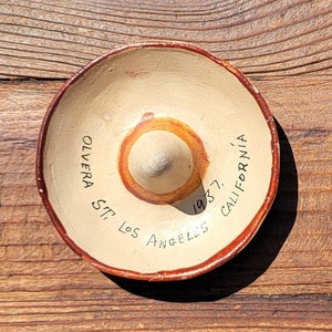 May include: A small, round, terracotta-colored sombrero with a white interior and a red and white design. The sombrero has the text "OLVERA ST. LOS ANGELES 1957 CALIFORNIA" written on it.