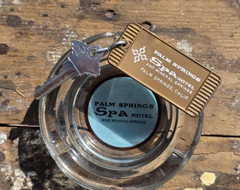 May include: A clear glass ashtray with a light blue center featuring the text "PALM SPRINGS Spa HOTEL AND MINERAL SPRINGS". A metal key and a gold-colored tag with the same text are also present.