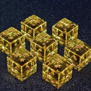 Puede incluir: Siete dados transparentes de color amarillo dorado con formas de estrella roja incrustadas. Los dados están dispuestos sobre una superficie oscura y brillante, creando un tema celestial. Cada dado tiene forma de cubo, con estrellas visibles en cada cara.