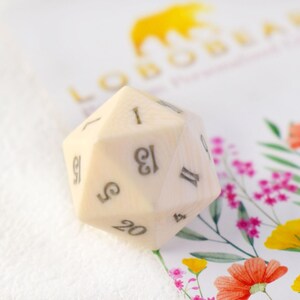 May include: A white 20-sided die with black numbers. The die is sitting on a white surface with a floral pattern.