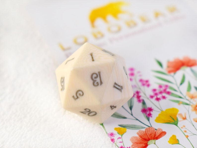 May include: A white 20-sided die with black numbers. The die is sitting on a white surface with a floral pattern.