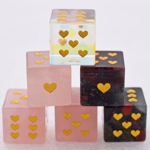 K&ouml;nnte beinhalten: Ein Satz von sechs herzf&ouml;rmigen W&uuml;rfeln, mit drei rosa W&uuml;rfeln und drei schwarzen W&uuml;rfeln. Die W&uuml;rfel sind aus klarem, schimmerndem Glas mit goldenen Herzen auf jeder Seite.