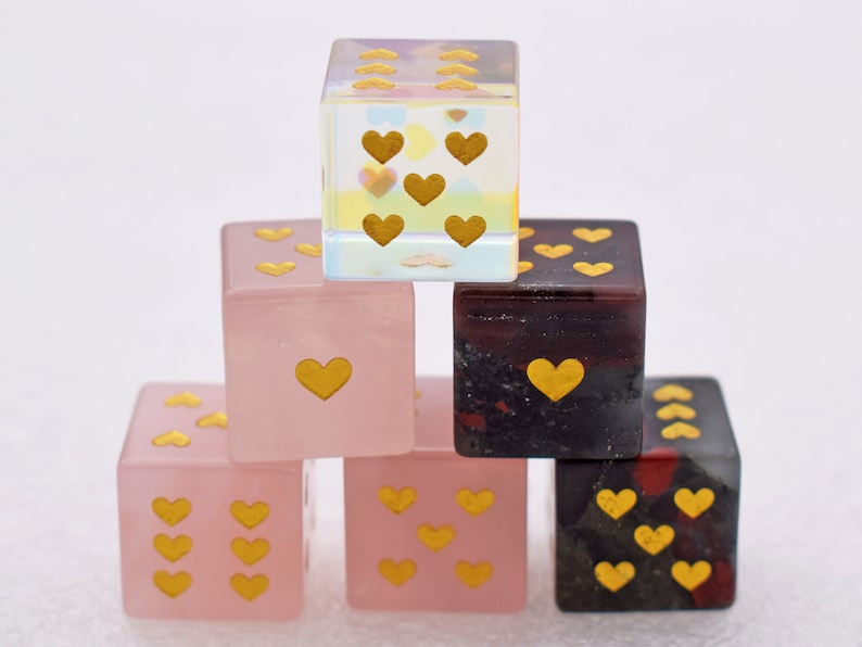 K&ouml;nnte beinhalten: Ein Satz von sechs herzf&ouml;rmigen W&uuml;rfeln, mit drei rosa W&uuml;rfeln und drei schwarzen W&uuml;rfeln. Die W&uuml;rfel sind aus klarem, schimmerndem Glas mit goldenen Herzen auf jeder Seite.