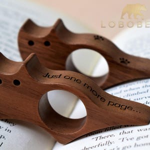 Puede incluir: Dos sujetap&aacute;ginas de libros de madera con forma de gato. Los sujetadores son marrones con una abertura circular para el dedo y el texto "Just one more page..." grabado en el lateral. Tambi&eacute;n son visibles huellas de patas.