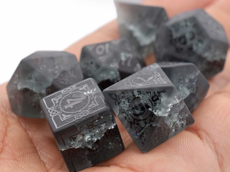 May include: A set of six black and grey dice with a clear, speckled finish. Each die has a different number from 1 to 6, with a decorative border around the number.