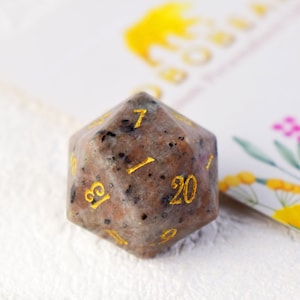 May include: A speckled brown and white 20-sided dice with gold numbers. The dice is made of natural stone and has a speckled appearance.