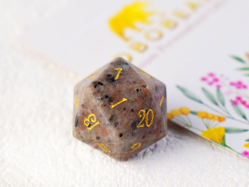 May include: A speckled brown and white 20-sided dice with gold numbers. The dice is made of natural stone and has a speckled appearance.