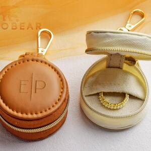 May include: Two round, zippered cases with gold-tone lobster clasps. One case is tan with the letters E|P embossed on the lid. The other case is cream-colored and open, revealing a beige interior lining and a gold-tone ring with small white beads.