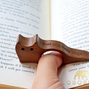 Puede incluir: Un sujetap&aacute;ginas de madera en forma de gato, con el texto "Just one more page..." grabado. El sujetap&aacute;ginas es marr&oacute;n y est&aacute; sobre las p&aacute;ginas de un libro.