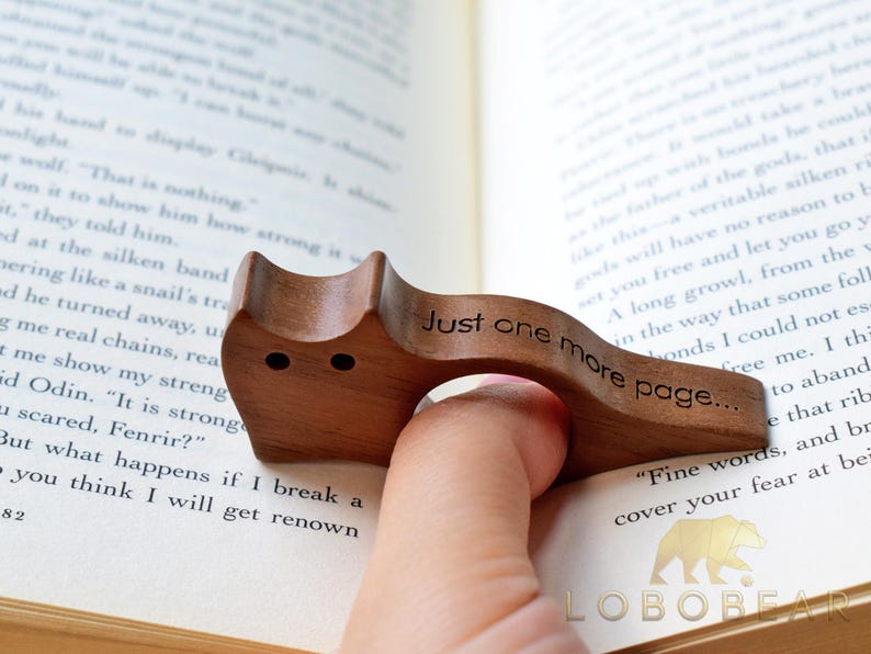 Puede incluir: Un sujetap&aacute;ginas de madera en forma de gato, con el texto "Just one more page..." grabado. El sujetap&aacute;ginas es marr&oacute;n y est&aacute; sobre las p&aacute;ginas de un libro.