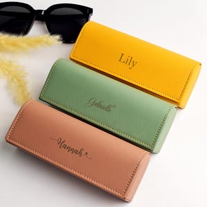 Könnte beinhalten: Drei personalisierte Brillenetuis in Gelb, Grün und Pfirsich. Jedes Etui hat einen eingravierten Namen: Lily, Gabrielle und Hannah. Schwarze Sonnenbrillen und dekoratives gelbes Gras befinden sich im Hintergrund.