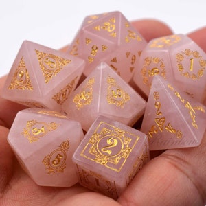 May include: A set of seven pink gemstone dice with gold numbers and intricate designs. The dice are in a hand, ready for a game.