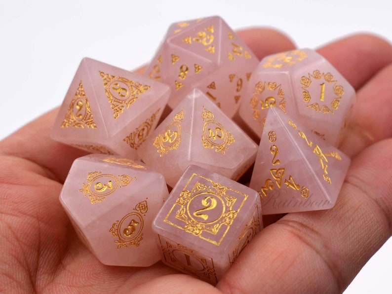 May include: A set of seven pink gemstone dice with gold numbers and intricate designs. The dice are in a hand, ready for a game.