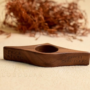 May include: A wooden book holder with a diamond shape and a circular hole. The holder is engraved with the words "book whore" and "spread those pages". The holder is brown and sits on a light-colored surface.