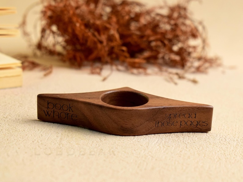 May include: A wooden book holder with a diamond shape and a circular hole. The holder is engraved with the words "book whore" and "spread those pages". The holder is brown and sits on a light-colored surface.