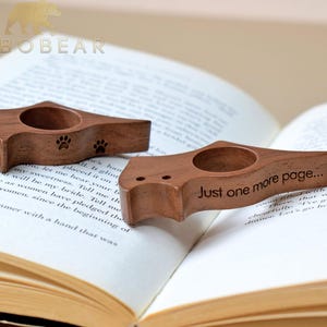 Puede incluir: Dos sujetalibros de madera con un dise&ntilde;o de huella de pata y el texto "Just one more page..." se muestran en un libro abierto. Los sujetalibros son de madera marr&oacute;n oscuro y tienen una abertura circular para el pulgar.