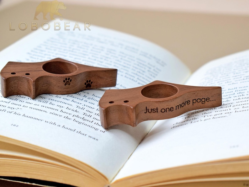 Puede incluir: Dos sujetalibros de madera con un dise&ntilde;o de huella de pata y el texto "Just one more page..." se muestran en un libro abierto. Los sujetalibros son de madera marr&oacute;n oscuro y tienen una abertura circular para el pulgar.