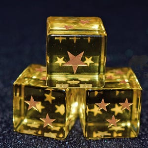 Könnte beinhalten: Drei transparente, goldgelbe Würfel mit braunen Sternmustern. Jeder Würfel hat einen großen Stern in der Mitte und kleinere Sterne an den Seiten. Die Würfel sind gestapelt, mit einem dunklen, glitzernden Hintergrund.