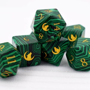Puede incluir: Un conjunto de siete dados poli&eacute;dricos verdes y negros, con n&uacute;meros dorados y un s&iacute;mbolo de lobo y luna creciente. Los dados, con efecto veteado, se muestran sobre un fondo blanco, ideales para juegos de mesa.