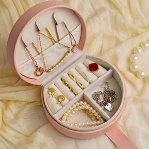 K&ouml;nnte beinhalten: Eine rosa, runde Schmuckschatulle, ge&ouml;ffnet, um verschiedene Halsketten, Ringe und Ohrringe zu pr&auml;sentieren. Der Schmuck umfasst Gold- und Ros&eacute;goldst&uuml;cke, eine Perlenkette und einen roten quadratischen Ring. Ein Perlenarmband ist ebenfalls sichtbar.