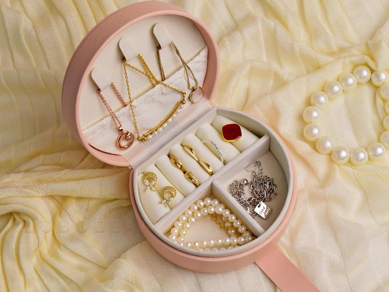 K&ouml;nnte beinhalten: Eine rosa, runde Schmuckschatulle, ge&ouml;ffnet, um verschiedene Halsketten, Ringe und Ohrringe zu pr&auml;sentieren. Der Schmuck umfasst Gold- und Ros&eacute;goldst&uuml;cke, eine Perlenkette und einen roten quadratischen Ring. Ein Perlenarmband ist ebenfalls sichtbar.