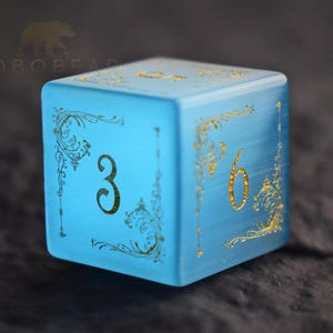 Peut inclure: Un d&eacute; bleu clair de forme carr&eacute;e, orn&eacute; de chiffres dor&eacute;s et de motifs d&eacute;coratifs. Les chiffres 3 et 6 sont visibles sur des faces adjacentes. Le d&eacute; a un aspect givr&eacute; et se d&eacute;tache sur un fond sombre.