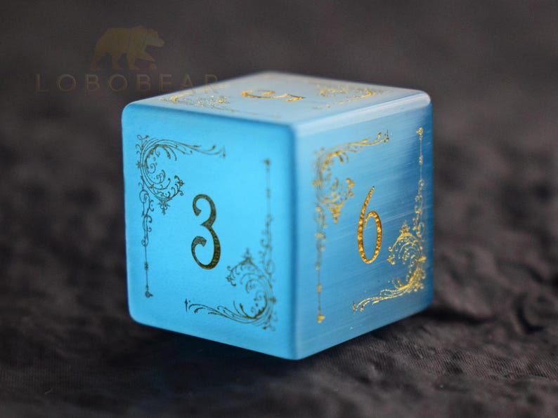 Peut inclure: Un d&eacute; bleu clair de forme carr&eacute;e, orn&eacute; de chiffres dor&eacute;s et de motifs d&eacute;coratifs. Les chiffres 3 et 6 sont visibles sur des faces adjacentes. Le d&eacute; a un aspect givr&eacute; et se d&eacute;tache sur un fond sombre.