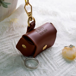 以下が含まれることがあります： ゴールドの留め具と「E&D 18.06.2024」というパーソナライズされた刻印が施された、茶色の革製のリングボックスです。